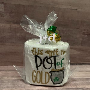 May include: A white toilet paper roll with gold and green embroidery that reads "This ain't no pot of gold". The roll is wrapped in clear plastic with a green and gold ribbon.