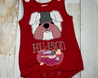 May include: Red sleeveless onesie with a white and black bulldog face embroidered on the front. The onesie has the name "Hudson" embroidered below the bulldog face.