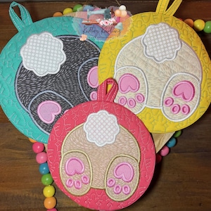 May include: Three circular quilted pot holders featuring embroidered bunny bottoms with fluffy white tails and pink paw pads. The pot holders are teal, yellow, and pink, each with a hanging loop at the top. A logo with the text Krittrz Kreations is visible.