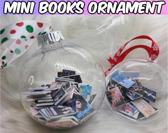 May include: Two clear glass ornaments filled with miniature books. The ornaments are decorated with red and white ribbon and snowflakes. The text on the books includes "The Hating Game" and "The Play."