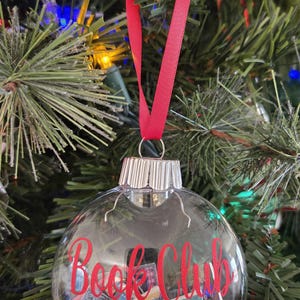 May include: A clear, spherical glass ornament featuring a silver metal cap and a red ribbon hanger. The exterior is printed with the red text "Book Club 2024," while the interior is filled with miniature rectangular book-shaped paper cutouts.