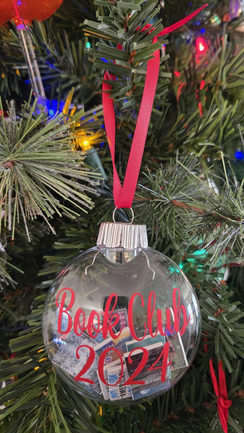 May include: A clear, spherical glass ornament featuring a silver metal cap and a red ribbon hanger. The exterior is printed with the red text "Book Club 2024," while the interior is filled with miniature rectangular book-shaped paper cutouts.