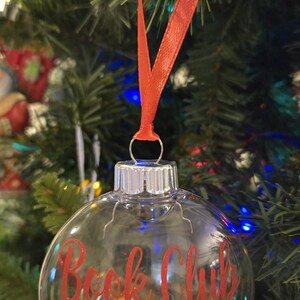 May include: A clear, spherical glass ornament hanging from a red ribbon. The exterior features the red text "Book Club 2025". Inside the transparent globe are several miniature book replicas with visible covers, creating a decorative holiday display.
