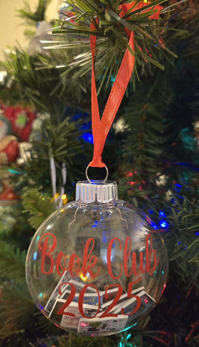 May include: A clear, spherical glass ornament hanging from a red ribbon. The exterior features the red text "Book Club 2025". Inside the transparent globe are several miniature book replicas with visible covers, creating a decorative holiday display.