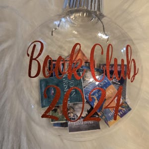 May include: A clear, spherical glass ornament featuring a red ribbon hanger and a silver cap. The exterior is decorated with red glittery text that reads Book Club 2024. Inside the ornament are several miniature rectangular book covers.