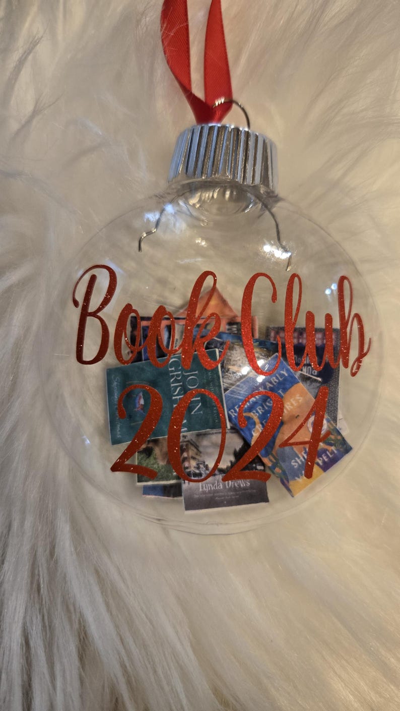 May include: A clear, spherical glass ornament featuring a red ribbon hanger and a silver cap. The exterior is decorated with red glittery text that reads Book Club 2024. Inside the ornament are several miniature rectangular book covers.