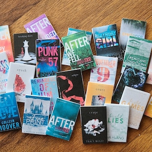 May include: A collection of 20 paperback books with various titles and authors. The books are arranged in a semi-circle on a wooden surface. The books are all different colors and have different designs on their covers.