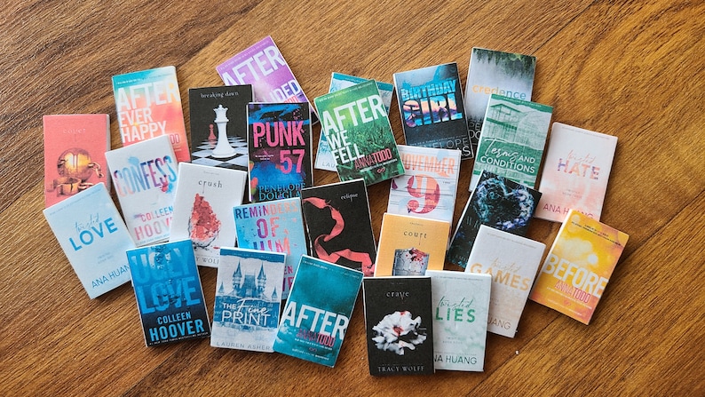 May include: A collection of 20 paperback books with various titles and authors. The books are arranged in a semi-circle on a wooden surface. The books are all different colors and have different designs on their covers.
