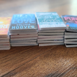 May include: A stack of paperback books with different covers. The covers feature various colors and designs, including a yellow cover with the word "BEFORE" on it, a blue cover with the words "It Ends With Us" and "Colleen Hoover" on it, a green cover with the words "Deal Offer" and "Lauren Asher" on it, and a pink cover with the words "Punk 57" and "Brooke Douglas" on it.