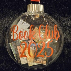 May include: A clear, spherical glass ornament filled with miniature book covers. The front of the ornament features the orange text "Book Club 2025" in a cursive font. A red ribbon loop is attached to the silver metal cap at the top of the ornament.