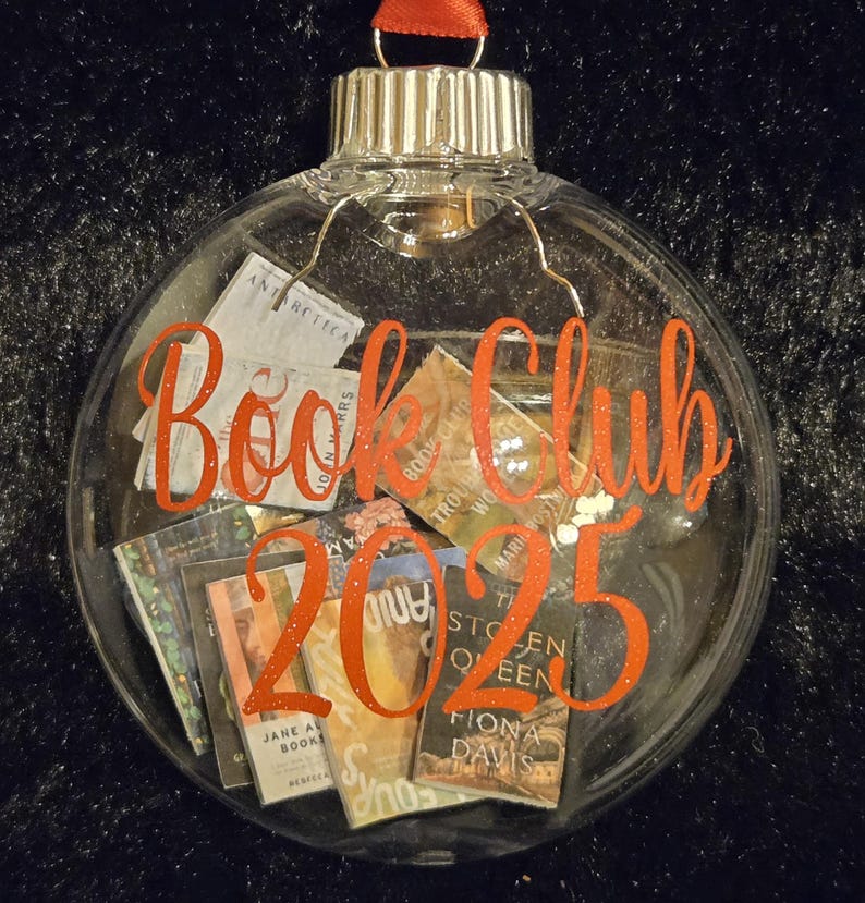 May include: A clear, spherical glass ornament filled with miniature book covers. The front of the ornament features the orange text "Book Club 2025" in a cursive font. A red ribbon loop is attached to the silver metal cap at the top of the ornament.