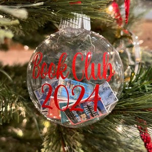 May include: A clear, spherical Christmas ornament filled with miniature book covers. The front of the ornament features red glittery text that reads "Book Club 2024" in a cursive script. The ornament is topped with a silver cap and a red ribbon.