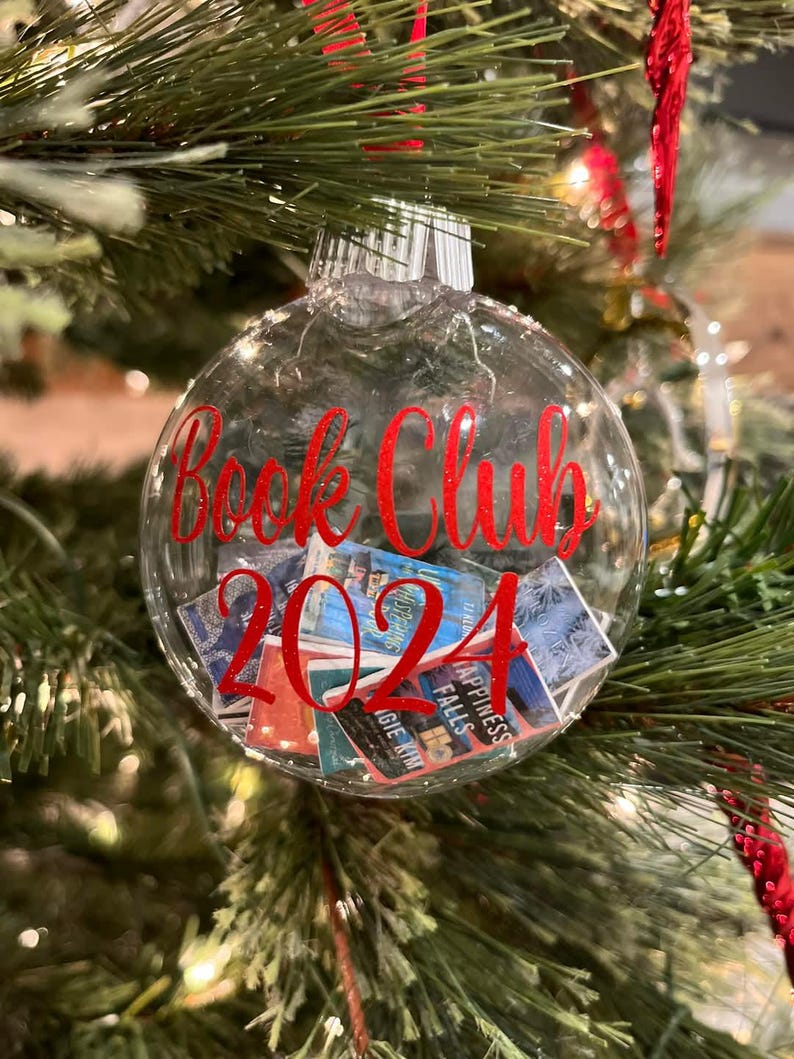 May include: A clear, spherical Christmas ornament filled with miniature book covers. The front of the ornament features red glittery text that reads "Book Club 2024" in a cursive script. The ornament is topped with a silver cap and a red ribbon.