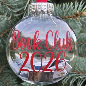 May include: A clear spherical glass ornament featuring a red ribbon hanger and a silver cap. The text "Book Club 2026" is printed in red on the front. Inside the glass sphere are several miniature books with visible covers and spines.