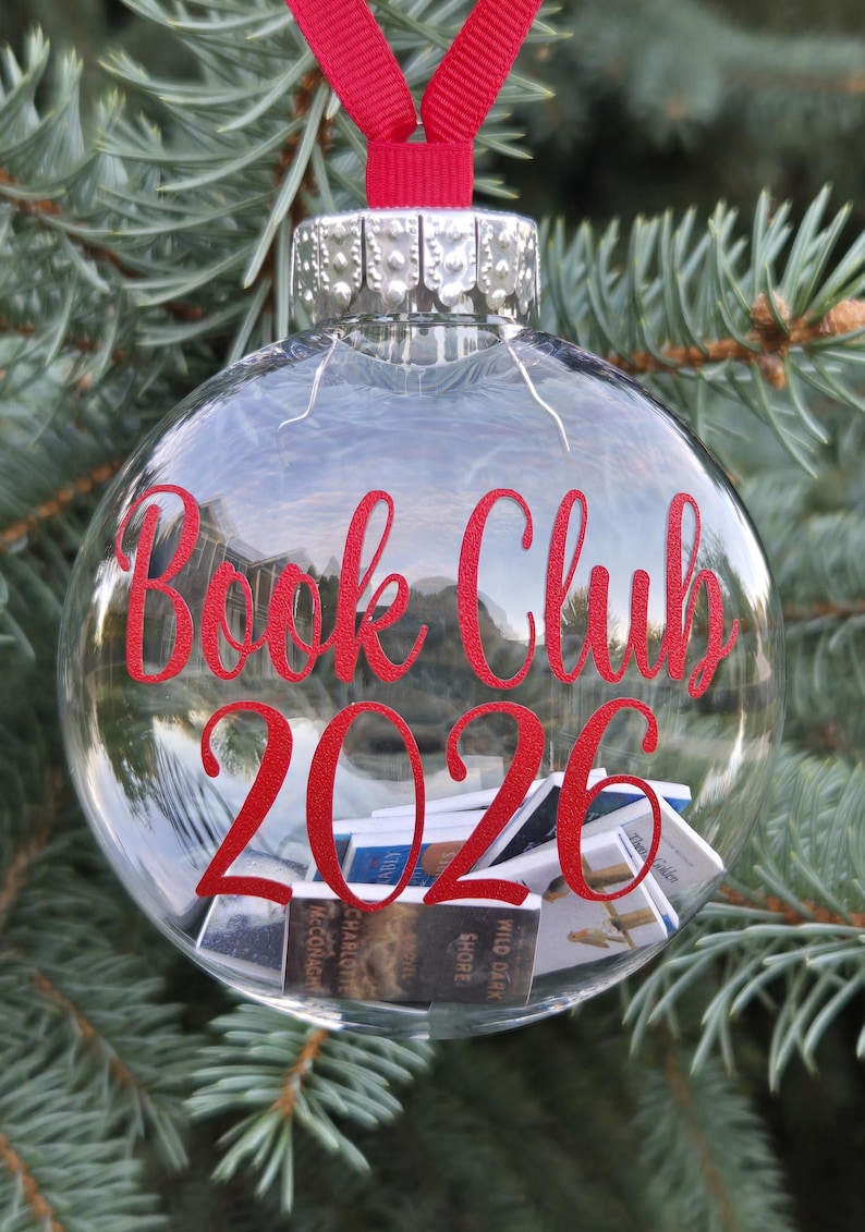 May include: A clear spherical glass ornament featuring a red ribbon hanger and a silver cap. The text "Book Club 2026" is printed in red on the front. Inside the glass sphere are several miniature books with visible covers and spines.