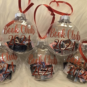 May include: Five clear, spherical glass ornaments, each featuring the red text "Book Club 2024" printed on the front. Inside each hollow ornament are miniature, colorful book covers. Each ornament is topped with a silver cap and a red ribbon loop.