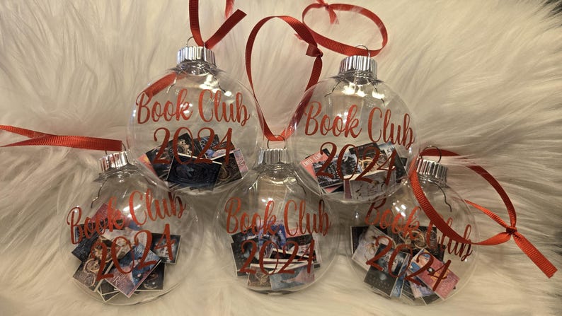 May include: Five clear, spherical glass ornaments, each featuring the red text "Book Club 2024" printed on the front. Inside each hollow ornament are miniature, colorful book covers. Each ornament is topped with a silver cap and a red ribbon loop.
