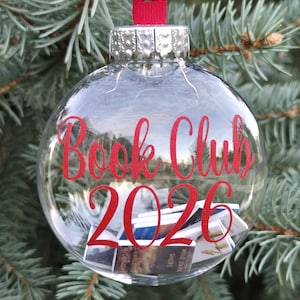 May include: A clear, spherical glass ornament featuring a red ribbon hanger and a silver cap. The text "Book Club 2026" is printed in red on the front. Inside the ornament are several miniature books with visible spines and covers.