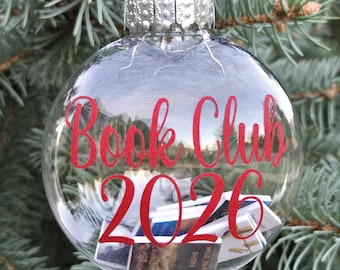 May include: A clear, spherical glass ornament featuring a red ribbon hanger and a silver cap. The text "Book Club 2026" is printed in red on the front. Inside the ornament are several miniature books with visible spines and covers.