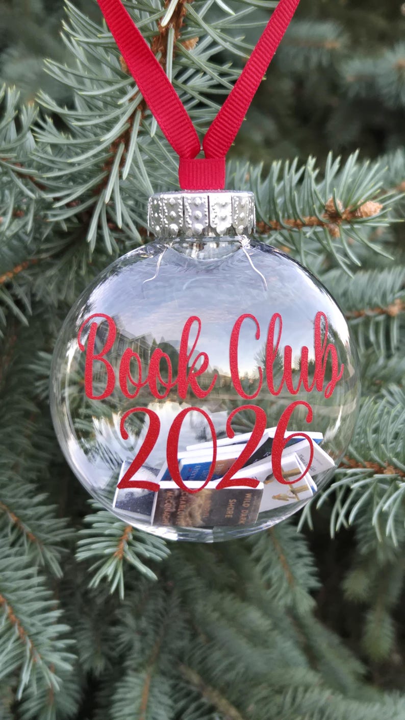 May include: A clear, spherical glass ornament featuring a red ribbon hanger and a silver cap. The text "Book Club 2026" is printed in red on the front. Inside the ornament are several miniature books with visible spines and covers.