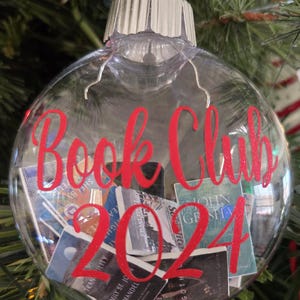 May include: A clear, spherical, hollow glass ornament featuring a red ribbon hanger and a silver cap. The exterior is printed with the red text "Book Club 2024." Inside the ornament are several miniature, rectangular book replicas.