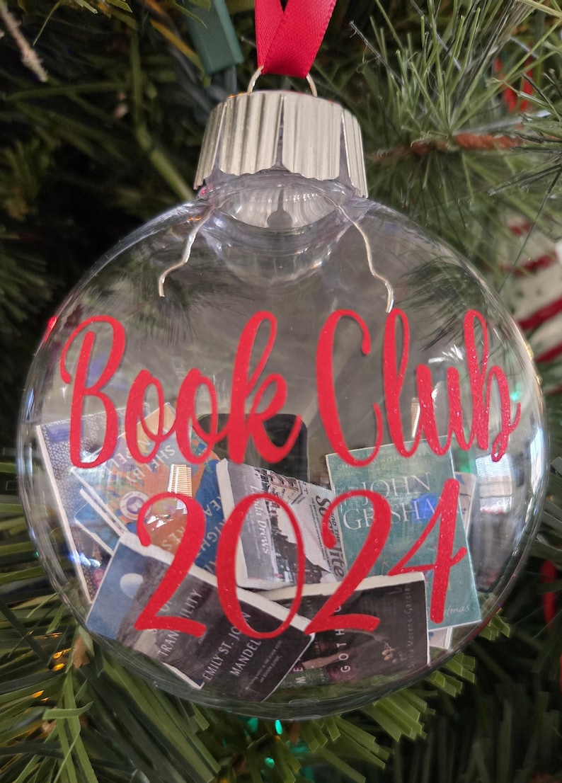May include: A clear, spherical, hollow glass ornament featuring a red ribbon hanger and a silver cap. The exterior is printed with the red text "Book Club 2024." Inside the ornament are several miniature, rectangular book replicas.