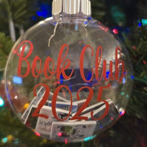 May include: A clear, spherical glass ornament featuring the red text "Book Club 2025" printed on the front. The interior of the hollow ornament is filled with miniature, rectangular book replicas with various colorful covers visible through the glass.