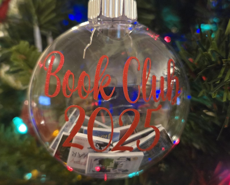 May include: A clear, spherical glass ornament featuring the red text "Book Club 2025" printed on the front. The interior of the hollow ornament is filled with miniature, rectangular book replicas with various colorful covers visible through the glass.