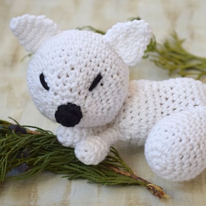 May include: White crocheted stuffed animal fox with black eyes and nose. The fox is lying down with its head resting on its paws.