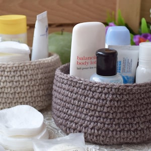 Puede incluir: Dos cestas de crochet llenas de diversos artículos de tocador, incluyendo loción, desinfectante de manos y discos de algodón. Las cestas son beige y marrón y están sobre una superficie blanca.