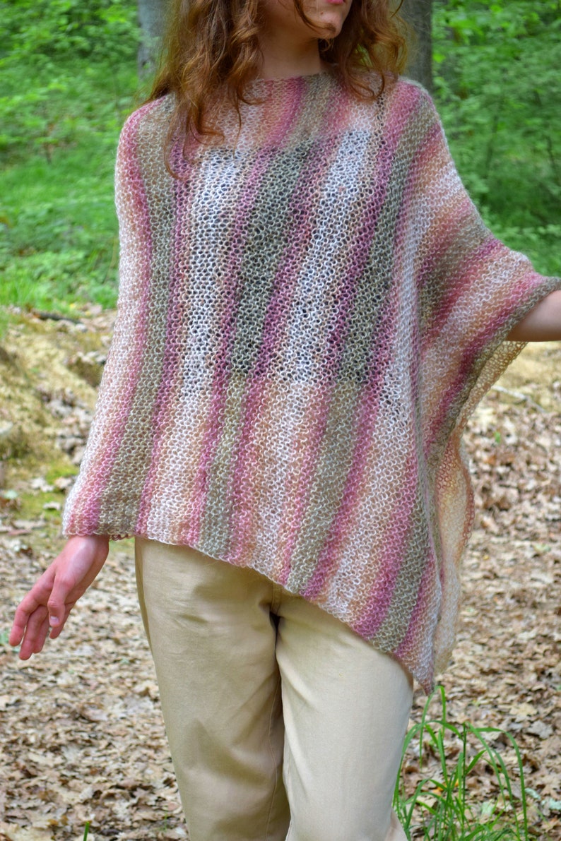 May include: A hand-knitted poncho in a striped pattern of pink, beige, and gray. The open weave garment has a relaxed fit and drapes over the shoulders. The poncho is a cozy, stylish accessory.