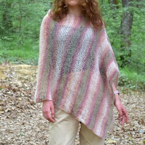 May include: A knitted poncho in shades of pink, white, and green. The poncho has a loose, asymmetrical design and is worn over a person's shoulders. The background features a forest setting with trees and foliage.