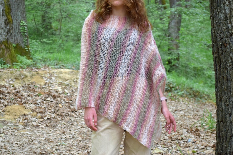 May include: A knitted poncho in shades of pink, white, and green. The poncho has a loose, asymmetrical design and is worn over a person's shoulders. The background features a forest setting with trees and foliage.