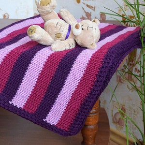 May include: A striped knitted baby blanket in shades of purple, pink, and burgundy. The blanket has a crocheted border and is lying on a wooden surface with a stuffed teddy bear on top.