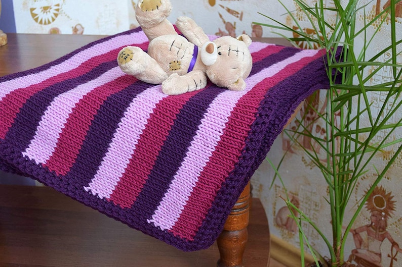 May include: A striped knitted baby blanket in shades of purple, pink, and burgundy. The blanket has a crocheted border and is lying on a wooden surface with a stuffed teddy bear on top.