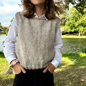 May include: A person wearing a light brown knitted vest with a white shirt and black pants. The vest has a scoop neck and is made of a textured yarn.