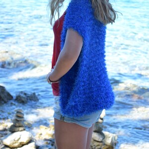 Puede incluir: Una mujer con un c&aacute;rdigan azul peludo sobre un top rojo sin mangas y pantalones cortos de mezclilla. Est&aacute; de pie en una playa rocosa con el oc&eacute;ano de fondo.