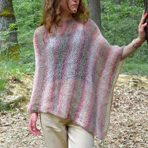 May include: A multi-colored, open-weave poncho in shades of pink, white, and green. The poncho has a loose, flowing design and is worn outdoors. The background features a forest setting with trees and foliage.