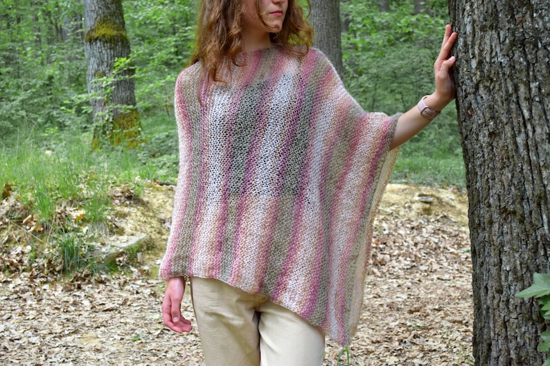 May include: A multi-colored, open-weave poncho in shades of pink, white, and green. The poncho has a loose, flowing design and is worn outdoors. The background features a forest setting with trees and foliage.