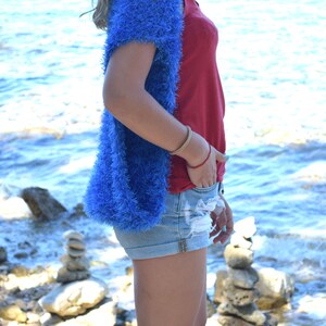 Puede incluir: Una mujer lleva un c&aacute;rdigan azul peludo sobre un top rojo sin mangas y pantalones cortos de mezclilla. Est&aacute; de pie en una playa rocosa con el oc&eacute;ano de fondo.