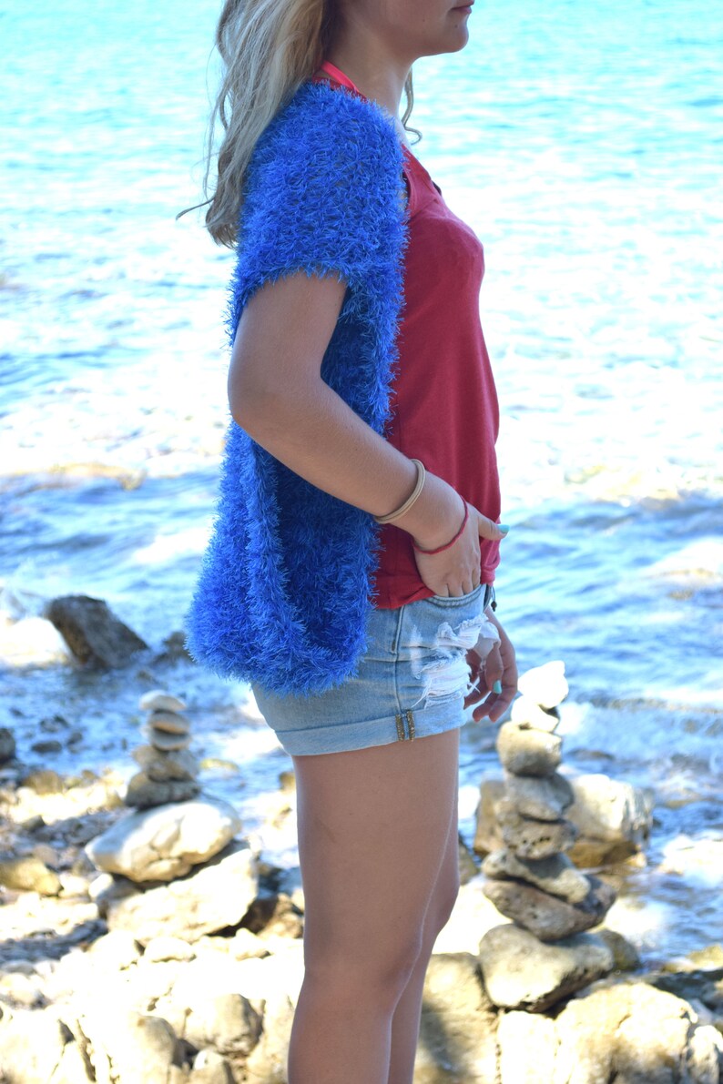 Puede incluir: Una mujer lleva un c&aacute;rdigan azul peludo sobre un top rojo sin mangas y pantalones cortos de mezclilla. Est&aacute; de pie en una playa rocosa con el oc&eacute;ano de fondo.