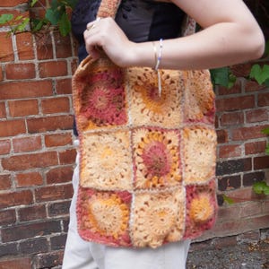 Könnte beinhalten: Eine handgefertigte Häkeltasche im Patchwork-Design. Die Tasche hat Quadrate in Pfirsich-, Orange- und Burgundertönen mit passendem Trageriemen. Die Tasche wird vor einer Backsteinmauer gehalten.