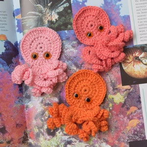 May include: Three crocheted octopus plushies with googly eyes. The octopus are pink, orange, and red. The plushies are sitting on a book about sea life.