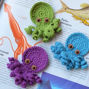 May include: Three crocheted octopus plushies in green, blue, and purple. Each octopus has eight tentacles and two large black eyes. The plushies are displayed on a book with text about sea creatures.