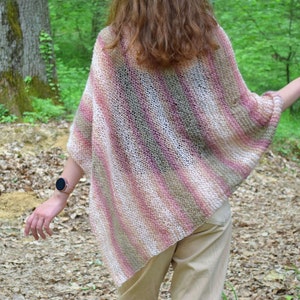 May include: A knitted poncho in shades of pink, white, and tan, with diagonal stripes. The open weave garment is worn outdoors, with a person's back to the camera. The background includes trees and foliage.