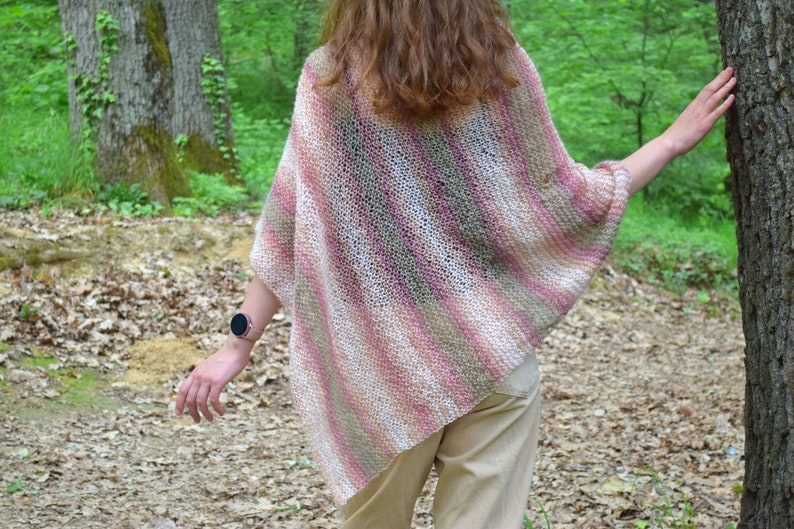 May include: A knitted poncho in shades of pink, white, and tan, with diagonal stripes. The open weave garment is worn outdoors, with a person's back to the camera. The background includes trees and foliage.