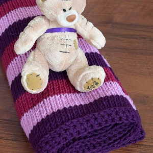 May include: A purple, pink, and burgundy striped knitted baby blanket with a beige teddy bear sitting on top. The teddy bear has a purple ribbon around its neck and the words "Me to You" on its foot.