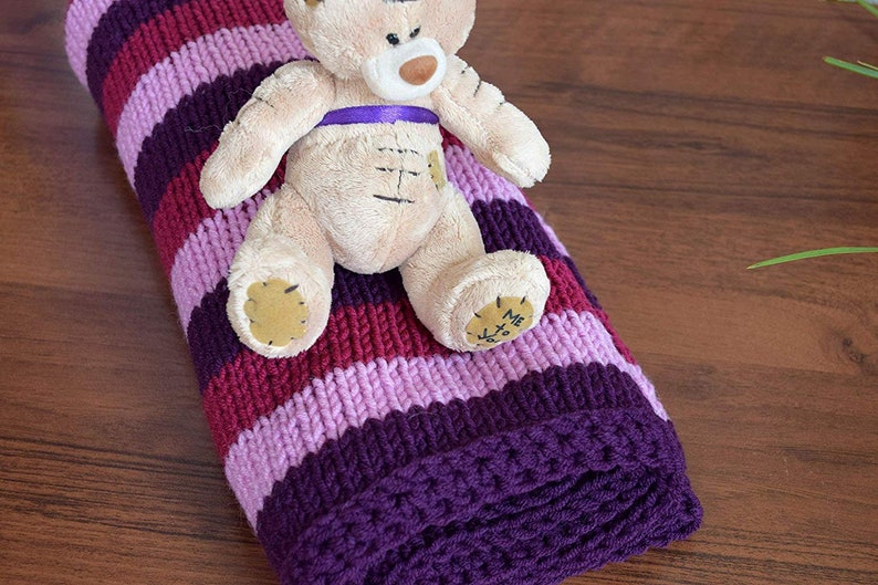 May include: A purple, pink, and burgundy striped knitted baby blanket with a beige teddy bear sitting on top. The teddy bear has a purple ribbon around its neck and the words "Me to You" on its foot.