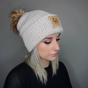 May include: A white knit beanie with a faux fur pom pom and a brown leather patch with a skull design.