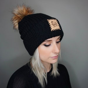 May include: Black knit beanie with a faux fur pom pom and a brown leather patch with a skull and crossbones design.